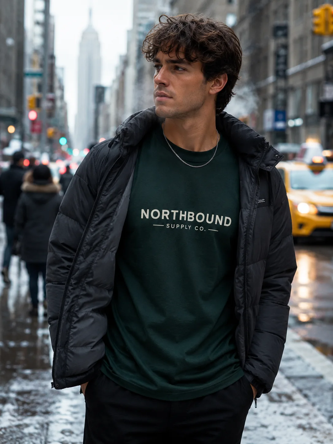 Model wearing a dark green logo T-shirt under an open winter jacket on a blurred New York street.