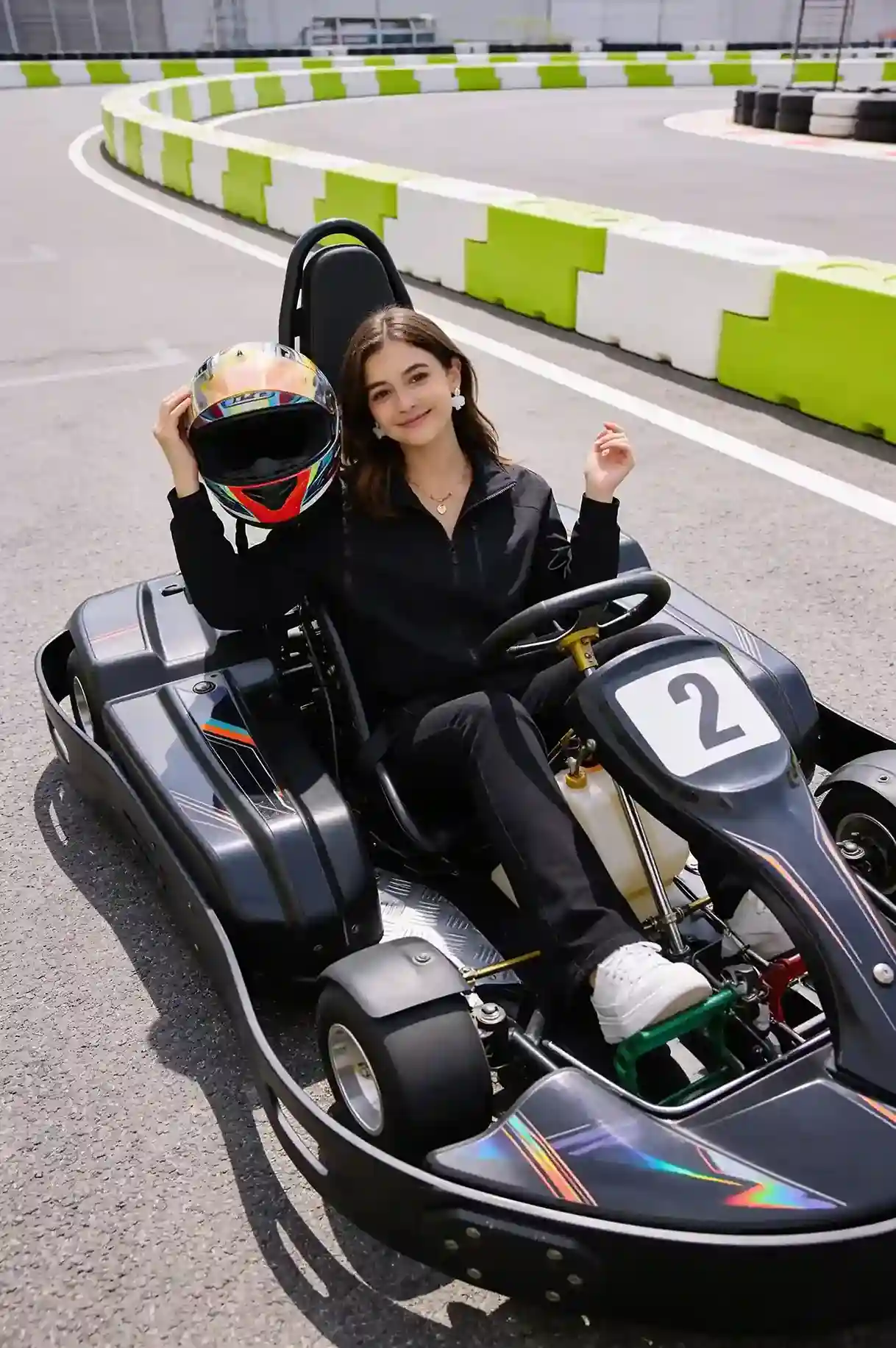 Person sitting in a racing go-kart while holding a helmet at an outdoor karting circuit with green and white barriers