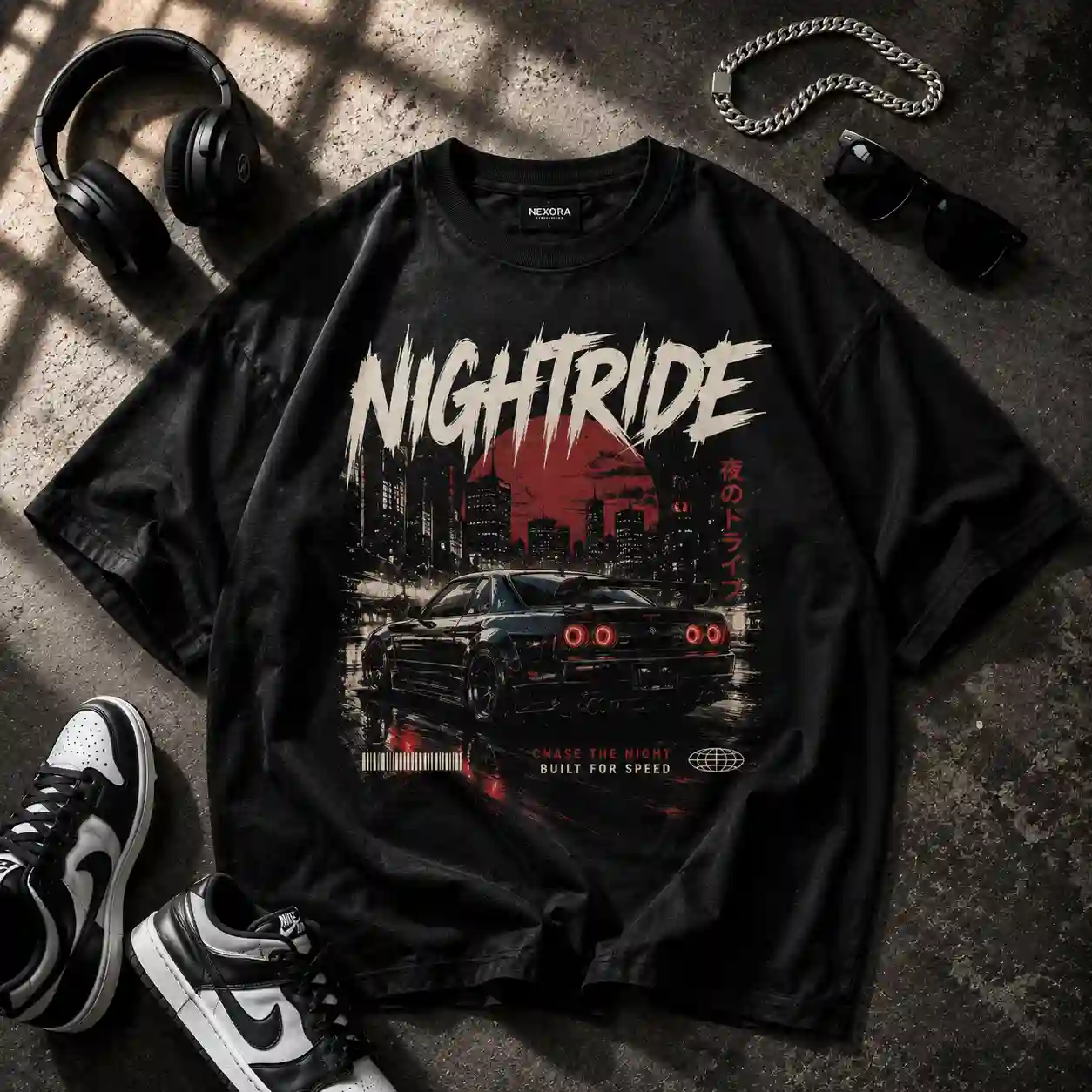 Black oversized streetwear T-shirt with a bold car graphic, styled with headphones, chain, sunglasses, and sneakers on a dark concrete background.