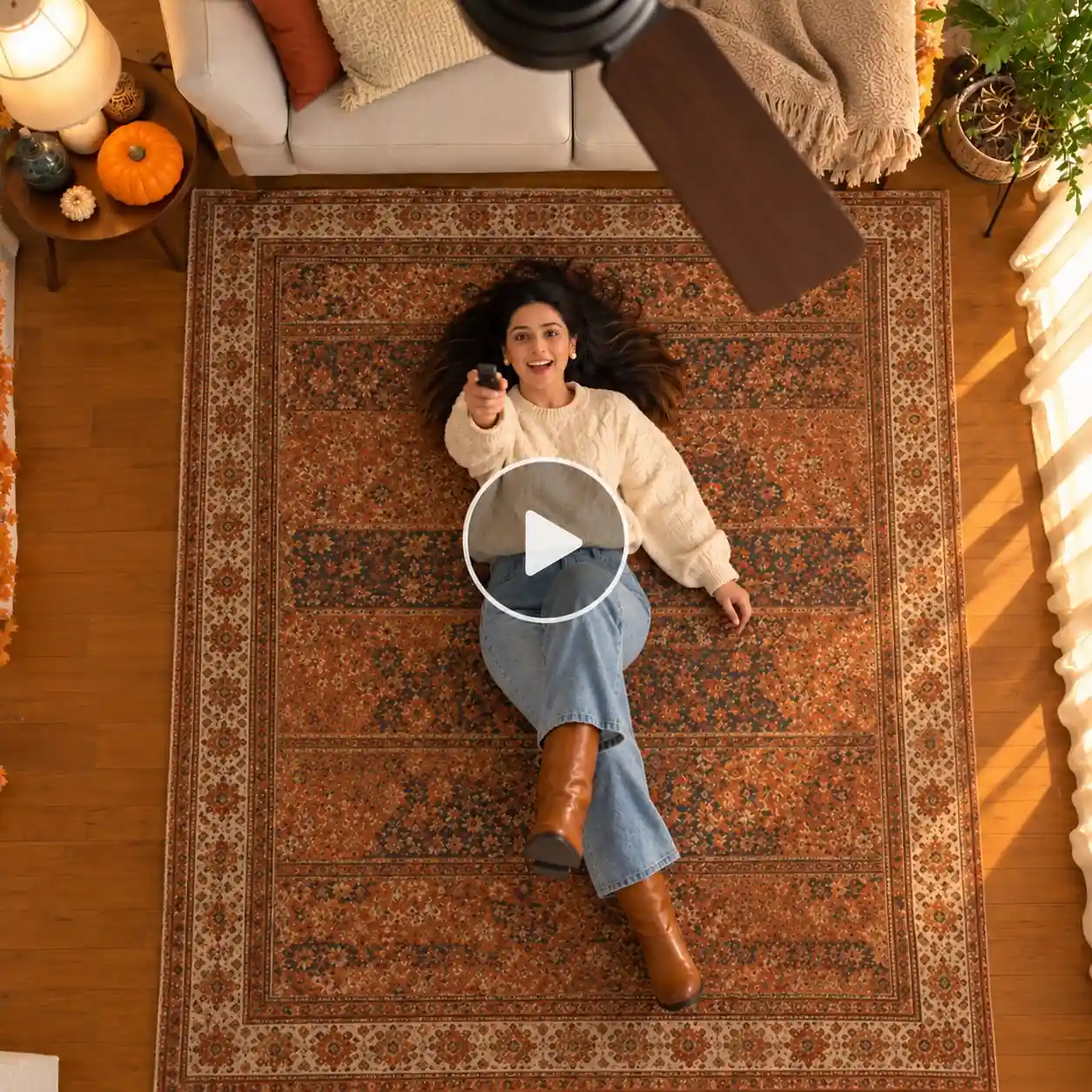 Cozy autumn ceiling fan trend with a woman lying on a rug, holding a remote, in a cream sweater and jeans.