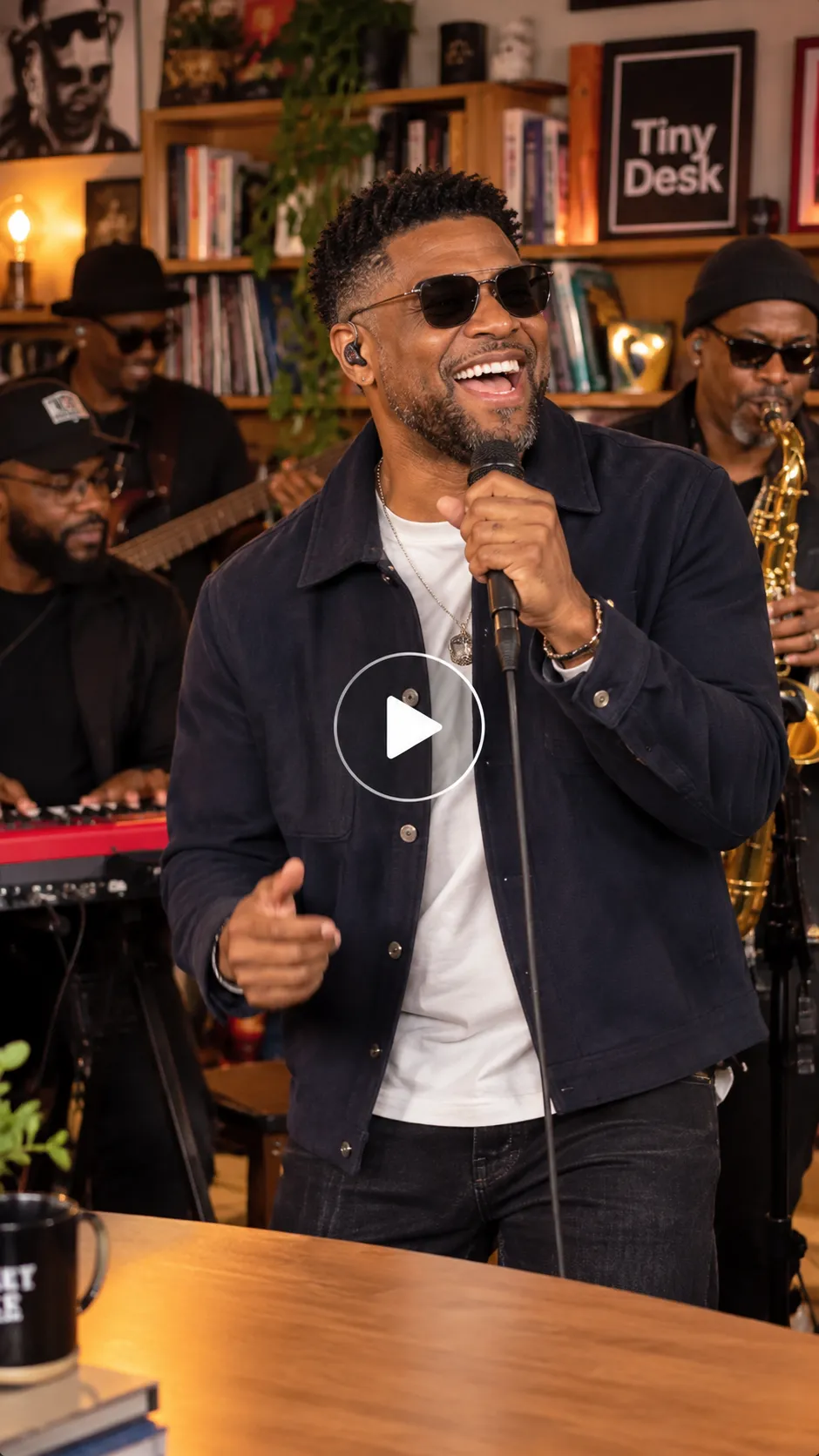 Realistic Tiny Desk AI video scene featuring a laughing lead singer with a microphone, a saxophonist, a red keyboard, warm lighting, bookshelves, and a transparent play button.