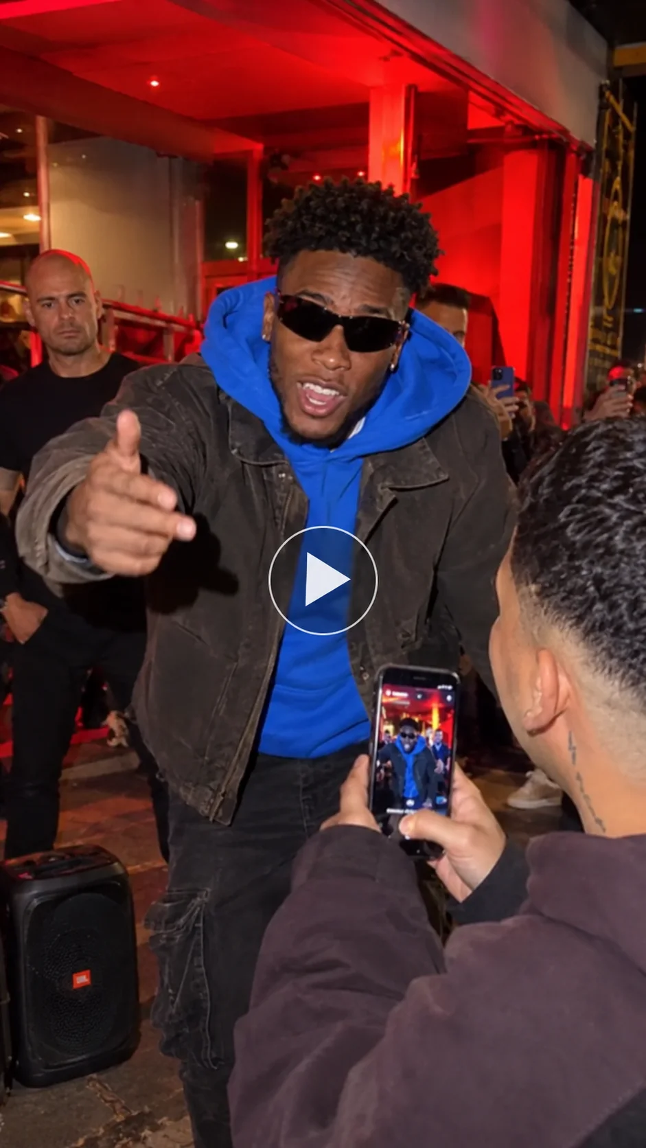 A man in a blue hoodie performs a street rap under red neon lights as a person records him on a phone, with a transparent play button in the center.